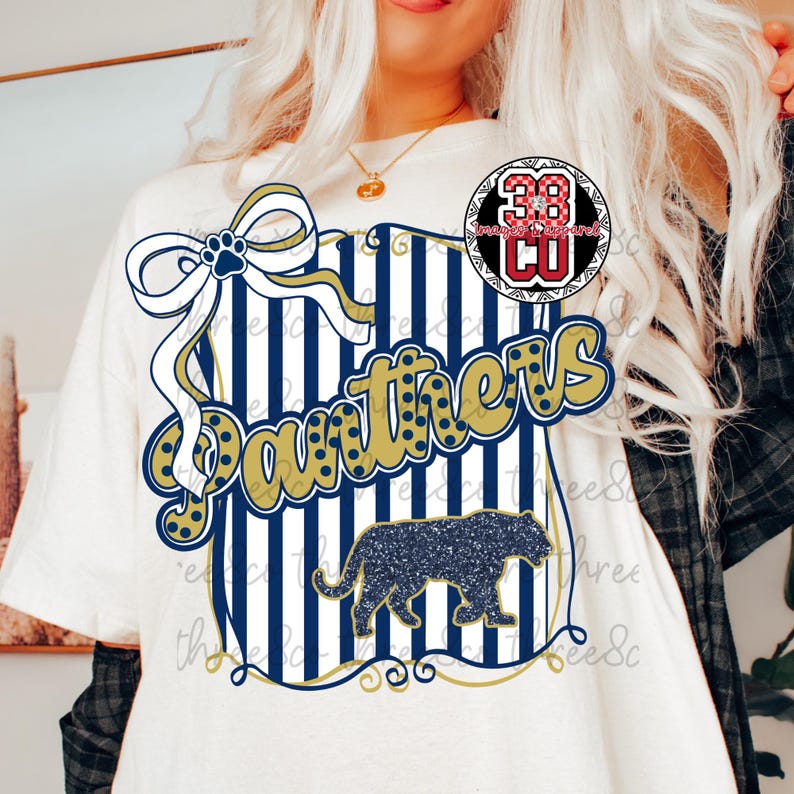 Puede incluir: Camiseta blanca con un dise&ntilde;o que presenta la palabra "Panthers" en dorado, una silueta de pantera azul marino y un lazo con una huella de pata. El dise&ntilde;o incluye rayas verticales azul marino y blancas.