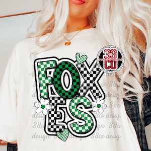 Puede incluir: Camiseta color crema con la palabra "FOXES" en un diseño a cuadros verdes y negros. El diseño incluye flores blancas y acentos de corazón verde. Un logotipo circular es visible en la parte superior derecha.