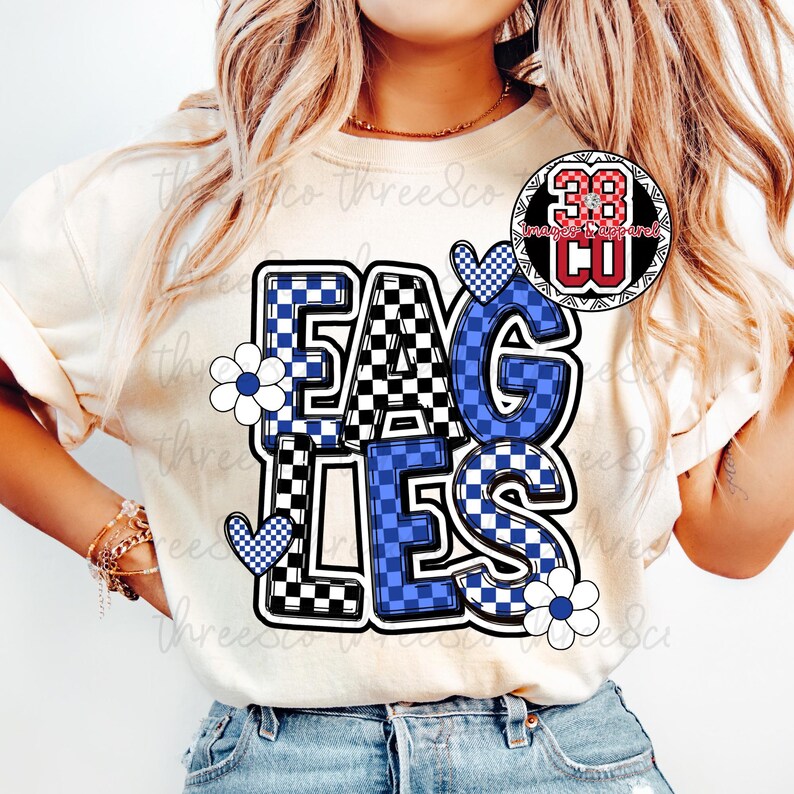 Puede incluir: Camiseta color crema con la palabra "EAGLES" en un dise&ntilde;o audaz a cuadros. Las letras alternan entre patrones de cuadros blancos y negros, y azules y blancos. Flores blancas y corazones a cuadros azules acent&uacute;an el dise&ntilde;o.