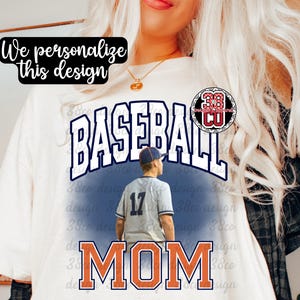 Può includere: T-shirt bianca con un design a tema baseball. La maglietta presenta la parola "BASEBALL" in grandi lettere blu navy e la parola "MOM" in arancione. È inclusa anche una grafica di un giocatore di baseball. Il testo sulla maglietta dice "We personalize this design."