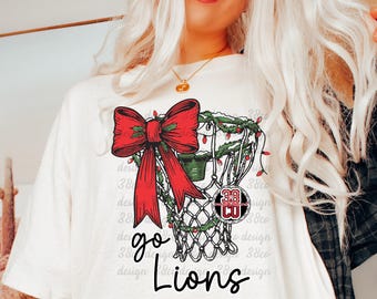May include: A cream-colored t-shirt featuring a graphic of a basketball hoop decorated with a large red bow, green garland, and red lights. The text "go Lions" is printed in black below the hoop, with a circular "38 CO" logo nearby.