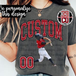 May include: Gray t-shirt with a baseball design. The word "CUSTOM" is in large red letters, with a baseball player graphic. The number "00" is at the bottom, and the text "We personalize this design" is at the top.