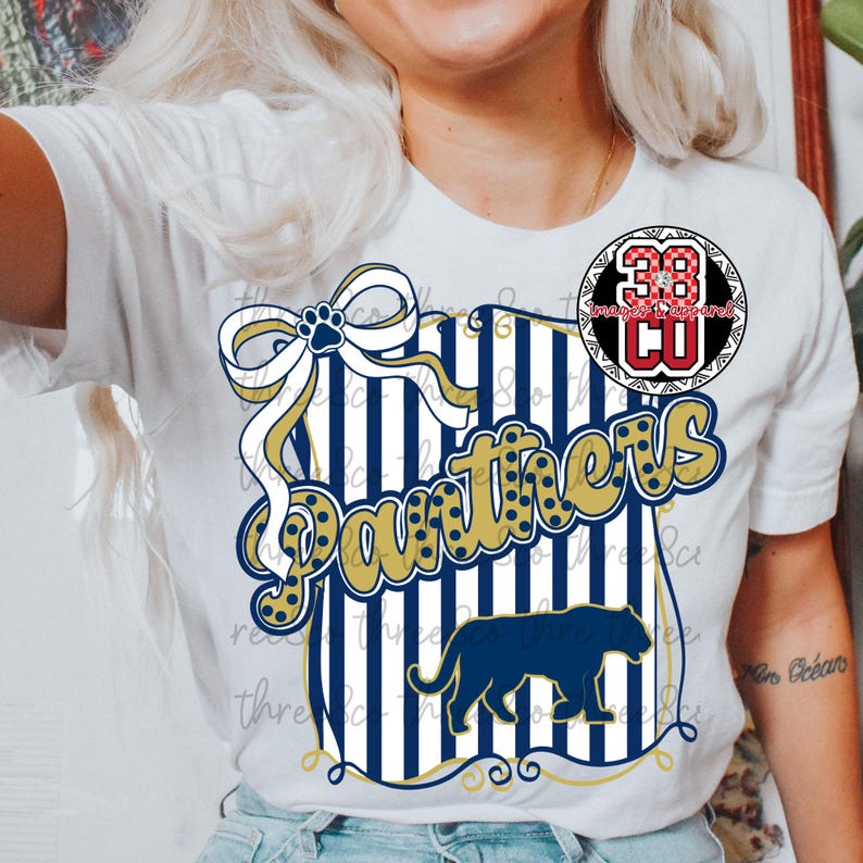 Puede incluir: Camiseta blanca con un dise&ntilde;o azul marino y dorado. El dise&ntilde;o incluye la palabra "Panthers", un lazo, rayas y una silueta de pantera. Tambi&eacute;n es visible un logotipo circular con el n&uacute;mero 38.