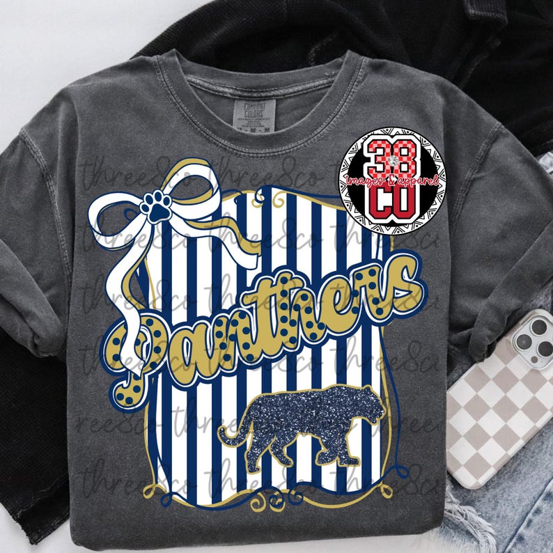 Puede incluir: Camiseta de manga larga gris oscuro con un dise&ntilde;o gr&aacute;fico. El dise&ntilde;o presenta la palabra "Panthers" en dorado y azul, una silueta de pantera y un lazo con una huella de pata. Tambi&eacute;n hay un logotipo circular con el n&uacute;mero 38.