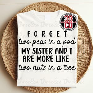 Op de afbeelding: Wit T-shirt met de zwarte tekst "FORGET two peas in a pod, MY SISTER AND I ARE MORE LIKE two nuts in a tree". Een cirkelvormig logo met de tekst "38 CU" staat rechtsboven. Het shirt ligt op een gevlochten mand.