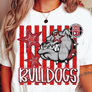May include: A white short-sleeve t-shirt featuring a graphic of a grey bulldog head with a spiked collar, red and white vertical stripes, two red stars, and the word BULLDOGS in white block letters with a black outline and a circular logo.