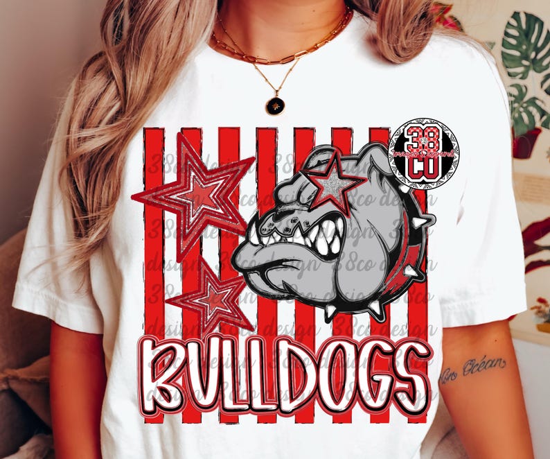 May include: A white short-sleeve t-shirt featuring a graphic of a grey bulldog head with a spiked collar, red and white vertical stripes, two red stars, and the word BULLDOGS in white block letters with a black outline and a circular logo.