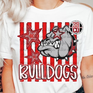 May include: A white short-sleeved t-shirt featuring a graphic of a grey bulldog head with a spiked collar, two red-outlined stars, and vertical red stripes. The word BULLDOGS is printed in large white letters with a red outline at the bottom.