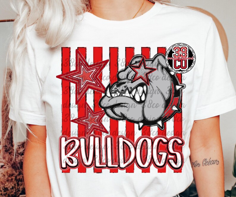 May include: A white short-sleeved t-shirt featuring a graphic of a grey bulldog head with a spiked collar, two red-outlined stars, and vertical red stripes. The word BULLDOGS is printed in large white letters with a red outline at the bottom.