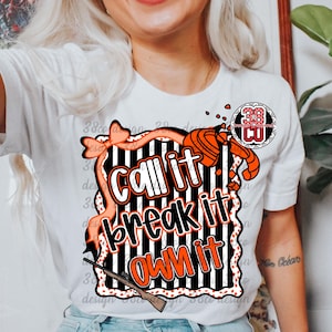 May include: White t-shirt featuring the text "Call it, Break it, Own it" in orange and white, with a black and white striped background. The design includes a bow, a shotgun, and a clay target.