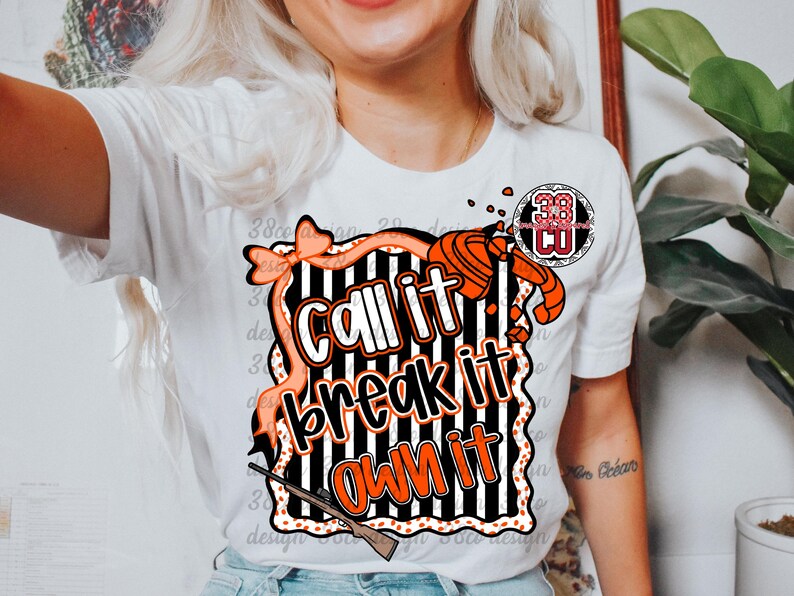 May include: White t-shirt featuring the text "Call it, Break it, Own it" in orange and white, with a black and white striped background. The design includes a bow, a shotgun, and a clay target.