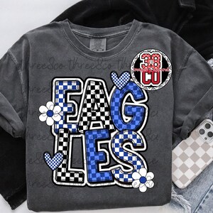 Puede incluir: Camiseta de manga larga gris oscuro con la palabra "EAGLES" en un patr&oacute;n de cuadros blanco, negro y azul. La camiseta tiene dise&ntilde;os florales y de coraz&oacute;n. Un parche circular con el n&uacute;mero "38" est&aacute; en la parte superior derecha.