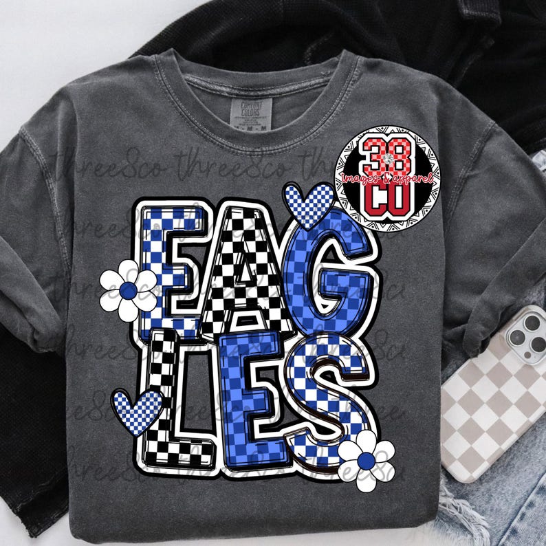 Puede incluir: Camiseta de manga larga gris oscuro con la palabra "EAGLES" en un patr&oacute;n de cuadros blanco, negro y azul. La camiseta tiene dise&ntilde;os florales y de coraz&oacute;n. Un parche circular con el n&uacute;mero "38" est&aacute; en la parte superior derecha.