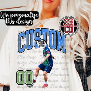 May include: Cream-colored t-shirt with the word "CUSTOM" in blue and black lettering. A soccer player graphic is featured, along with the number "00" in a green and black frame. The text "We personalize this design" is also present.