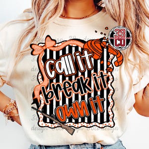 May include: Cream-colored t-shirt with a graphic design featuring the text "Call it, break it, own it" in white and orange. The design includes a shotgun, bow, and a black and white striped background.