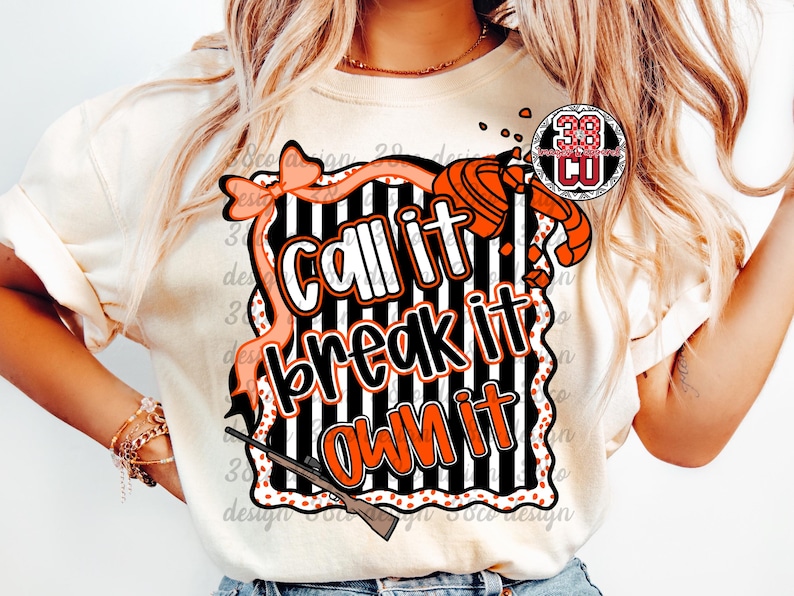 May include: Cream-colored t-shirt with a graphic design featuring the text "Call it, break it, own it" in white and orange. The design includes a shotgun, bow, and a black and white striped background.