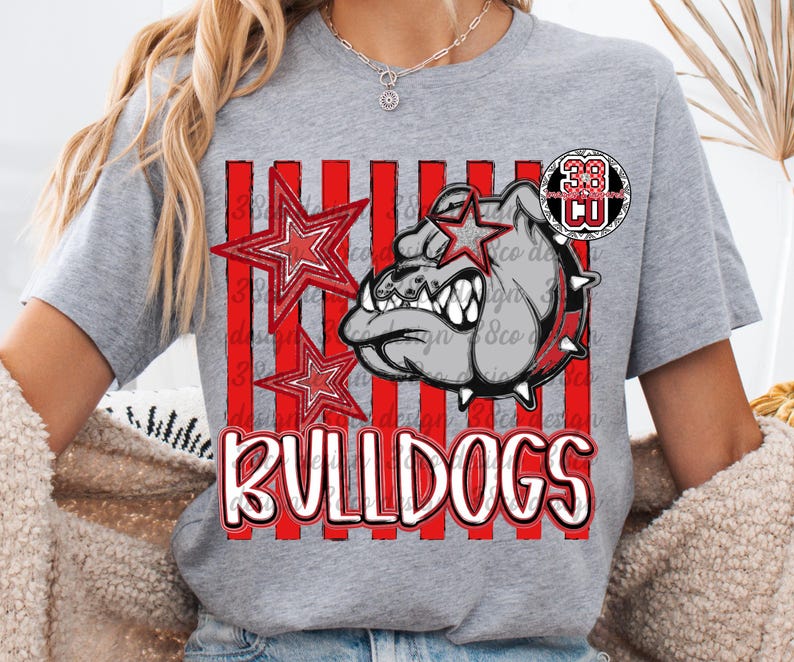 May include: A heather gray short-sleeve t-shirt featuring a graphic print of a bulldog wearing a spiked collar, two red stars, and vertical red stripes. The word BULLDOGS is printed in large white letters with a red outline at the bottom of the design.