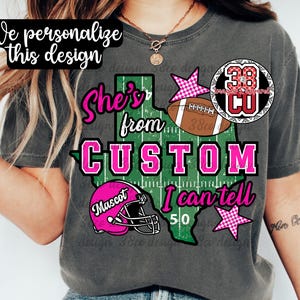 May include: A charcoal gray t-shirt featuring a graphic of the state of Texas designed like a football field. The text reads "She's from CUSTOM I can tell" and "Mascot". The design includes a brown football, a pink helmet, and pink checkered stars.