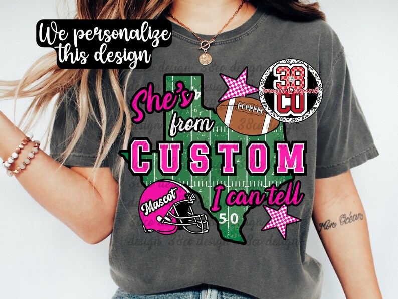 May include: A charcoal gray t-shirt featuring a graphic of the state of Texas designed like a football field. The text reads "She's from CUSTOM I can tell" and "Mascot". The design includes a brown football, a pink helmet, and pink checkered stars.
