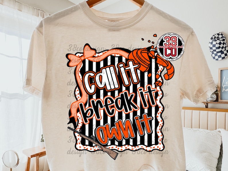 May include: Cream-colored t-shirt featuring a graphic design with the text "Call it, Break it, Own it" in white and orange. The design includes a bow, a shotgun, and a target, with black and white stripes.