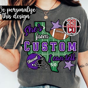 May include: A charcoal gray t-shirt featuring a graphic of the state of Texas with a football field pattern. The text reads "She's from CUSTOM I can tell" with a purple football helmet labeled "Mascot", a brown football, and purple checkered stars.