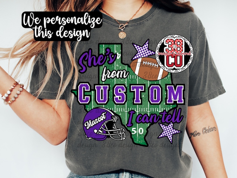 May include: A charcoal gray t-shirt featuring a graphic of the state of Texas with a football field pattern. The text reads "She's from CUSTOM I can tell" with a purple football helmet labeled "Mascot", a brown football, and purple checkered stars.