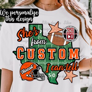 May include: A white t-shirt featuring a graphic of the state of Texas in green with white yard lines. The design includes the text "She's from CUSTOM I can tell", an orange football helmet, a brown football, and two orange and white checkered stars.