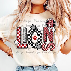 以下が含まれることがあります： 大胆なチェック柄で「LIONS」の文字がプリントされたクリーム色のTシャツ。文字は赤、黒、白で、星のアクセントが付いています。「38 CU」の円形エンブレムも表示されています。