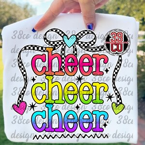 May include: White t-shirt with the word "cheer" in three lines, each in a different color gradient. The design includes a checkered bow, hearts, and stars. The shirt is held up by a hand.