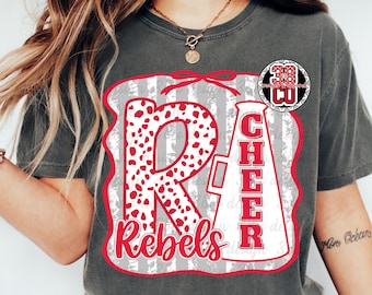 May include: A charcoal gray t-shirt featuring a graphic design with a large red and white dalmatian-spotted letter R, a white megaphone with the word CHEER in red letters, and the word Rebels in red script, all on a gray striped background.