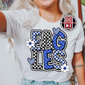 Puede incluir: Camiseta blanca con la palabra "EAGLES" en letras a cuadros azules y negros. El dise&ntilde;o incluye flores y corazones azules y blancos. Un logotipo circular es visible en la esquina superior derecha.
