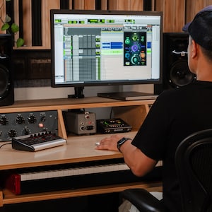 May include: A professional music production workstation featuring a computer monitor displaying digital audio software with a plugin interface, a MIDI keyboard controller, audio interface hardware, and studio speakers on a wooden desk.