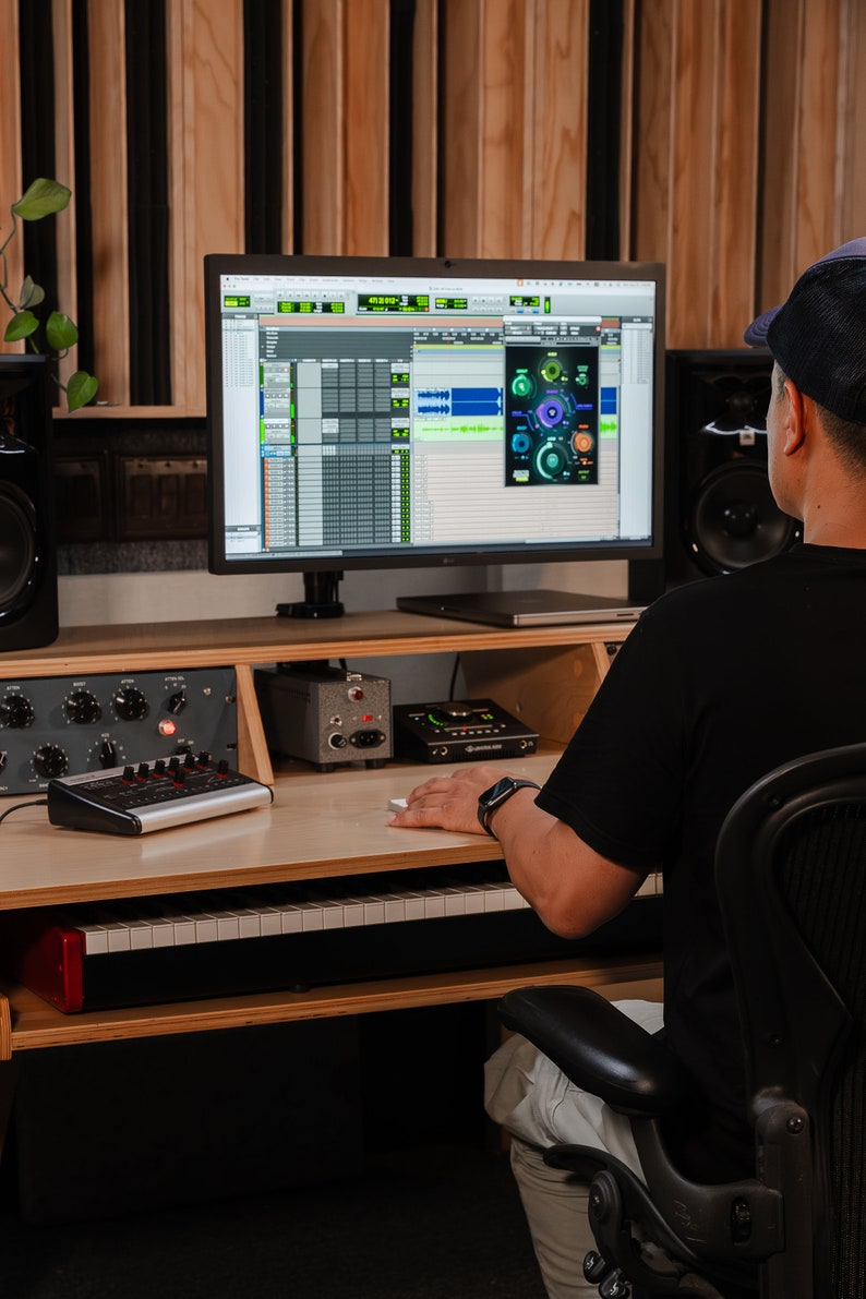 May include: A professional music production workstation featuring a computer monitor displaying digital audio software with a plugin interface, a MIDI keyboard controller, audio interface hardware, and studio speakers on a wooden desk.