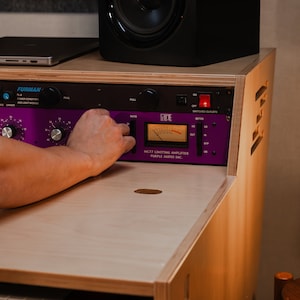 May include: A Purple Audio MC77 limiting amplifier with a purple faceplate, black knobs, and an analogue VU meter, mounted in a light-coloured wooden rack. Above it sits a black JBL studio monitor speaker and a Furman power conditioner unit.