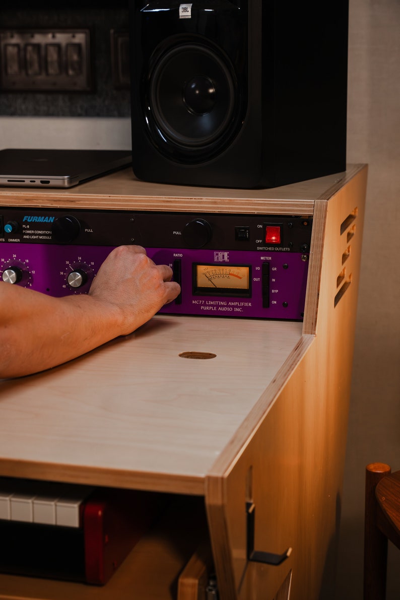 May include: A Purple Audio MC77 limiting amplifier with a purple faceplate, black knobs, and an analogue VU meter, mounted in a light-coloured wooden rack. Above it sits a black JBL studio monitor speaker and a Furman power conditioner unit.