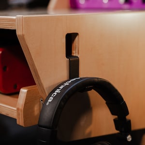 May include: A light-coloured plywood studio desk featuring a side-mounted black metal headphone hanger. The hanger is slotted into a rectangular cutout on the desk panel, supporting a pair of black over-ear headphones with the brand name visible.