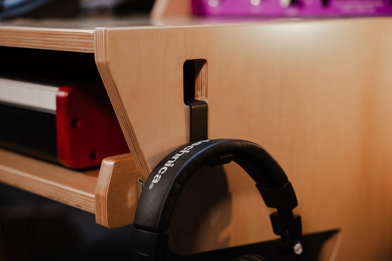 May include: A light-coloured plywood studio desk featuring a side-mounted black metal headphone hanger. The hanger is slotted into a rectangular cutout on the desk panel, supporting a pair of black over-ear headphones with the brand name visible.