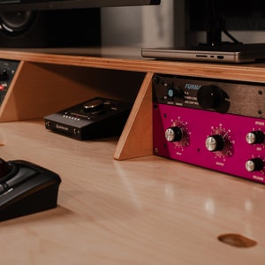 May include: A light-coloured wooden studio desk featuring a black trackball mouse, a black audio interface, and a rack-mounted audio processor with a bright magenta faceplate, silver knobs, and the text MC77 LIMITING AMPLIFIER PURPLE AUDIO.