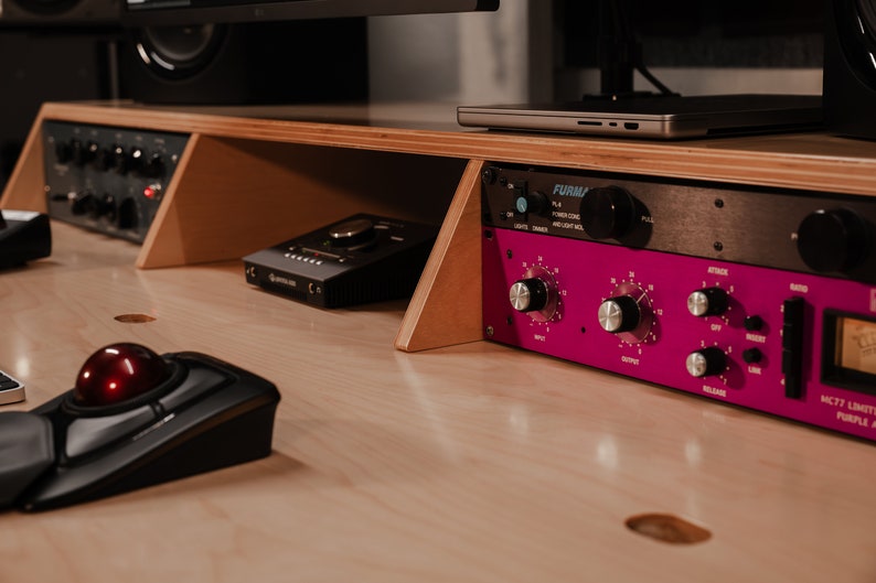 May include: A light-coloured wooden studio desk featuring a black trackball mouse, a black audio interface, and a rack-mounted audio processor with a bright magenta faceplate, silver knobs, and the text MC77 LIMITING AMPLIFIER PURPLE AUDIO.