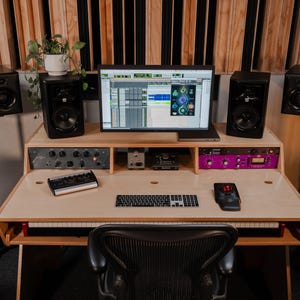 May include: A wooden studio desk featuring a computer monitor displaying audio software, a keyboard, a trackball mouse, and a small audio interface. The setup includes four black studio monitors on stands, rack-mounted gear, and a black office chair.