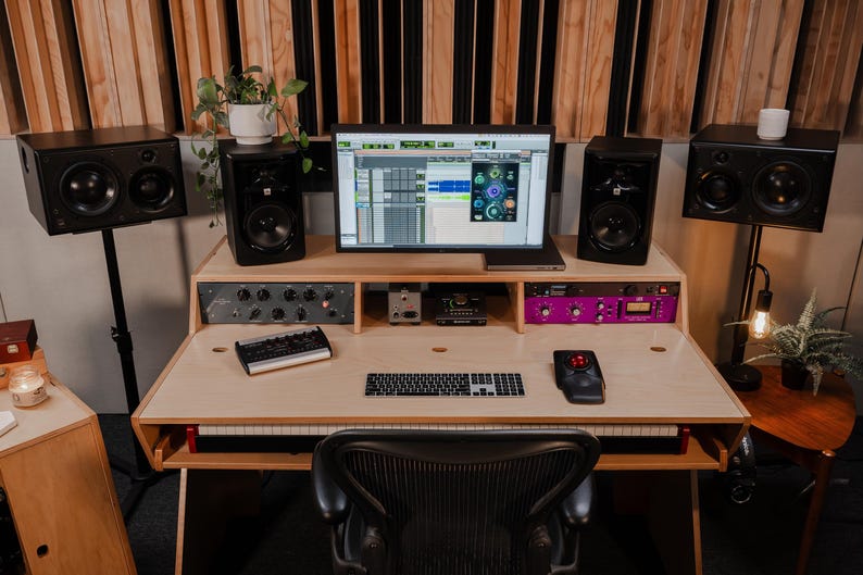 May include: A wooden studio desk featuring a computer monitor displaying audio software, a keyboard, a trackball mouse, and a small audio interface. The setup includes four black studio monitors on stands, rack-mounted gear, and a black office chair.