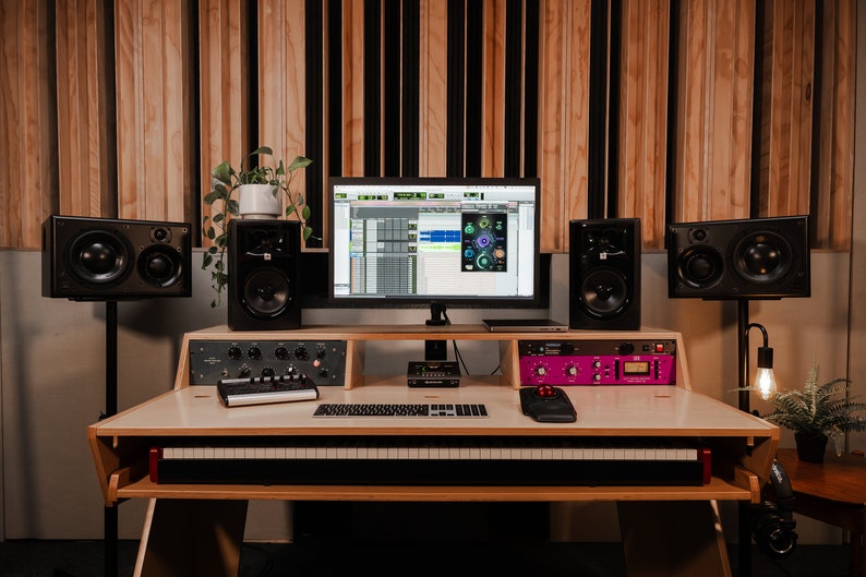 May include: A wooden studio desk featuring a built-in keyboard tray, a computer monitor displaying audio software, four black studio monitors on stands, a grey keyboard, a black mouse, and rack-mounted audio equipment with black and purple panels.