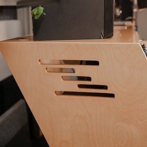 May include: A light-coloured wooden studio desk featuring a slanted side panel with four horizontal, staggered rectangular cutouts. The desk surface is smooth with a natural wood grain finish, and the side panel has a distinct angled profile.