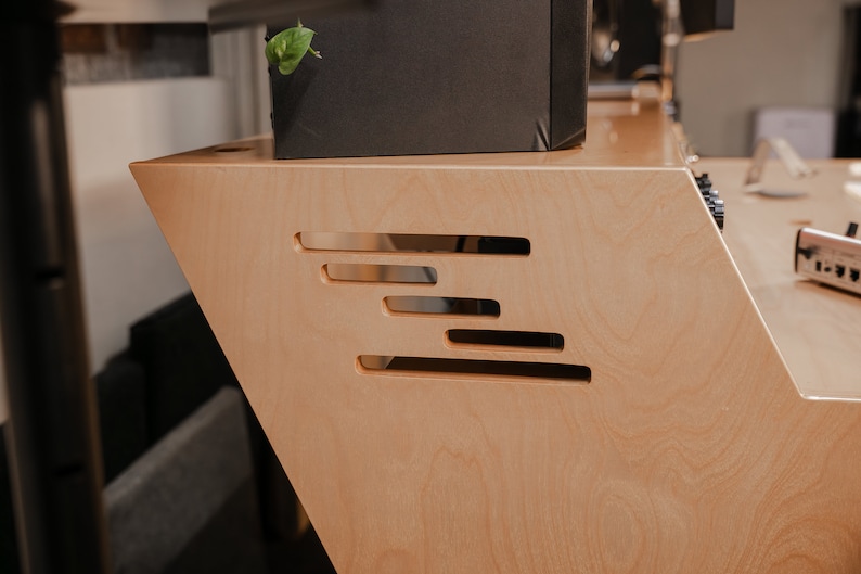 May include: A light-coloured wooden studio desk featuring a slanted side panel with four horizontal, staggered rectangular cutouts. The desk surface is smooth with a natural wood grain finish, and the side panel has a distinct angled profile.