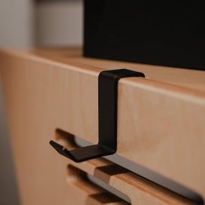 May include: A matte black metal hook with a flat, L-shaped design that clips over the edge of a light-coloured wooden surface. The hook features a vertical arm that extends downwards and a small, angled tab at the bottom to hold items securely.