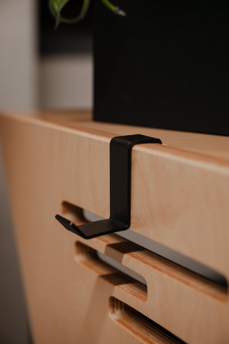 May include: A matte black metal hook with a flat, L-shaped design that clips over the edge of a light-coloured wooden surface. The hook features a vertical arm that extends downwards and a small, angled tab at the bottom to hold items securely.
