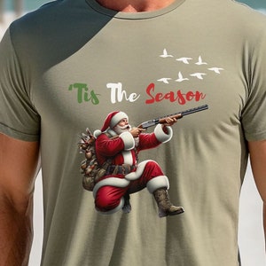 May include: Olive green t-shirt featuring Santa Claus aiming a shotgun, with the text "Tis The Season" above. Santa is depicted in his traditional red suit and boots, with a sack of gifts. White birds fly in the background.