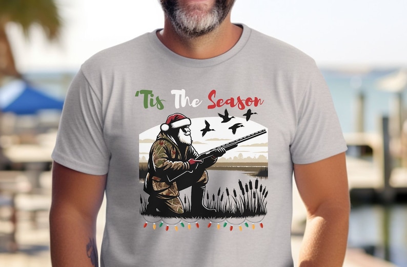 May include: A gray t-shirt with a Christmas-themed design featuring a Santa Claus character holding a shotgun and aiming at flying ducks. The text "Tis The Season" is printed above the image. A string of Christmas lights is printed below the image.