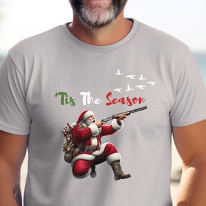 May include: Light gray t-shirt featuring a graphic of Santa Claus aiming a shotgun, with the text "'Tis The Season" in green and red. Above Santa, a flock of white birds flies across the sky. Santa is wearing a red suit and boots.