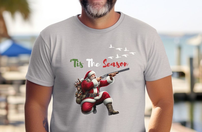 May include: Light gray t-shirt featuring a graphic of Santa Claus aiming a shotgun, with the text "'Tis The Season" in green and red. Above Santa, a flock of white birds flies across the sky. Santa is wearing a red suit and boots.